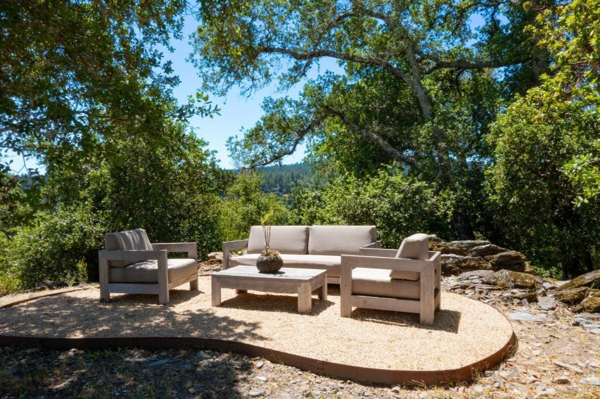 A seating area with a sofa and chairs is situated on a gravel surface under trees. 