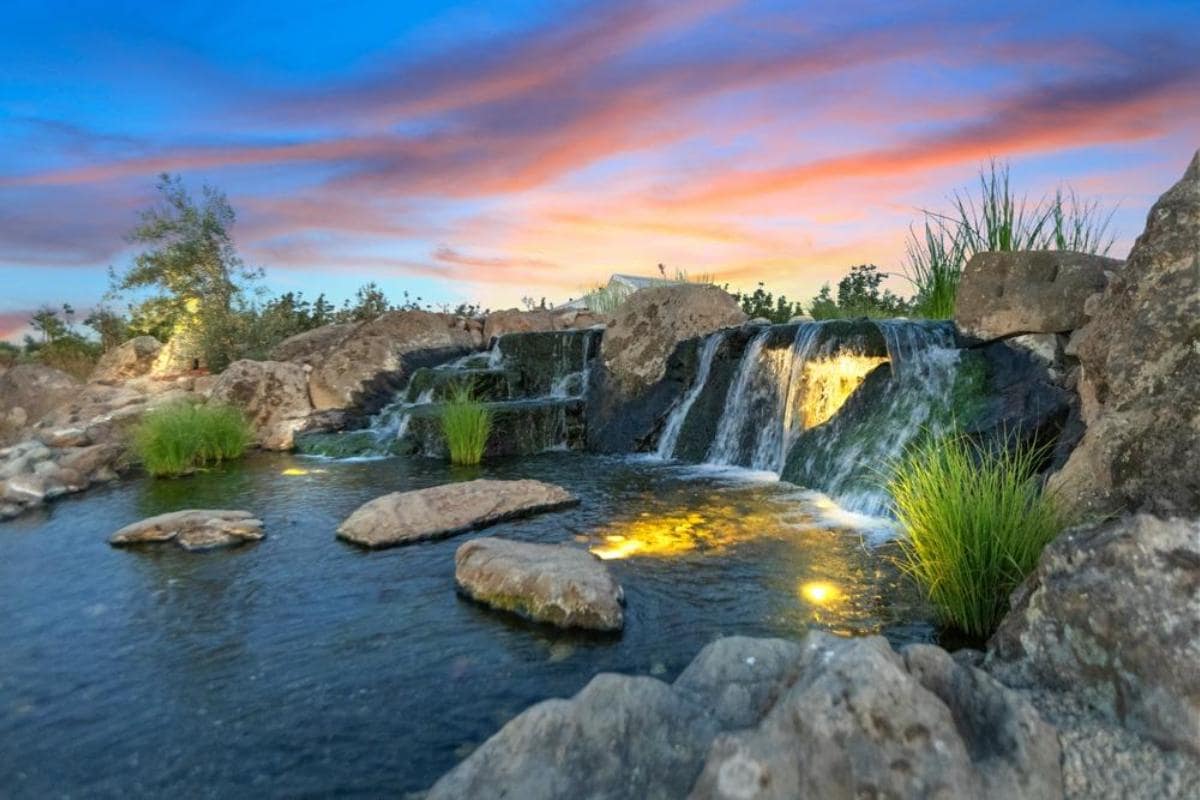 A beautiful waterfall cascading over rocks into a serene pond, illuminated by lights, with a colorful sunset in the background.