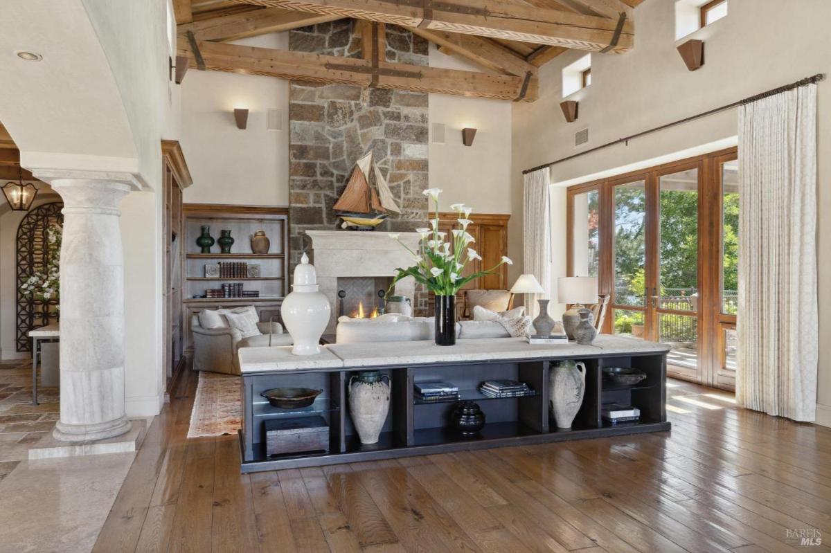 A living room with a stone fireplace, wooden beams, a central coffee table, and large windows leading to an outdoor area.