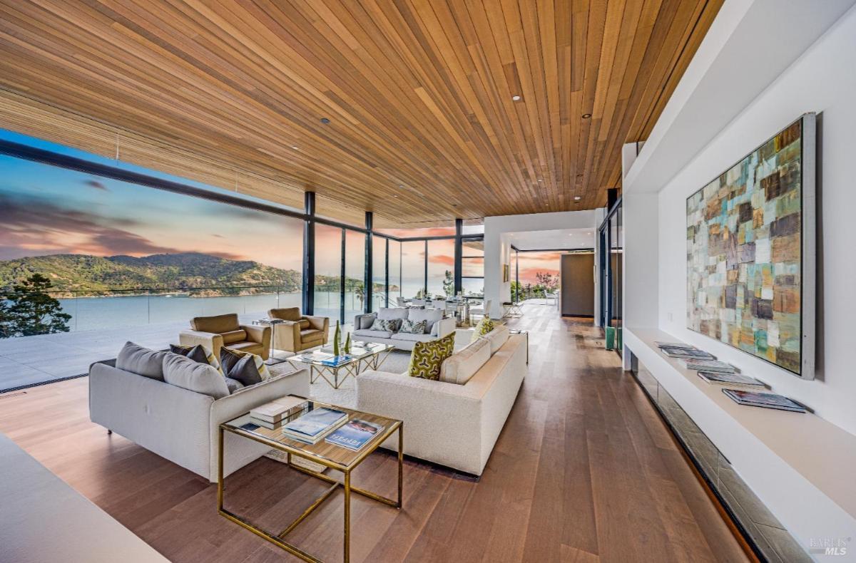 A spacious living area with floor-to-ceiling glass, a wood-panel ceiling, modern sofas, and a view at sunset.