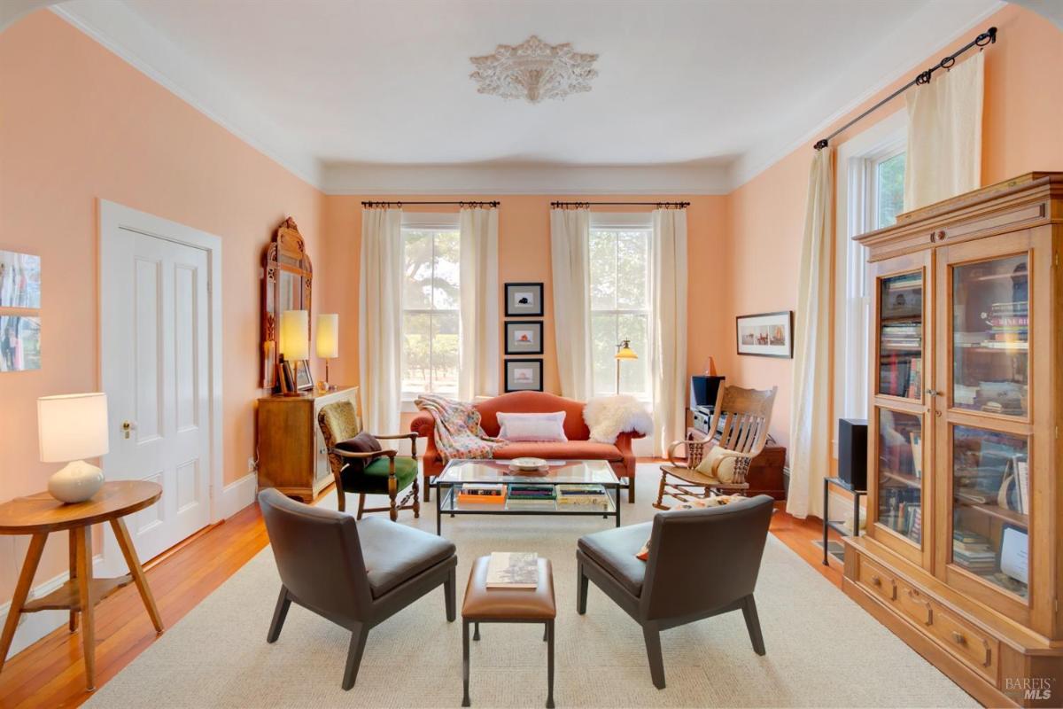 A living room with peach walls, large windows, and a mix of furniture including a sofa, rocking chair, and a wooden display cabinet.