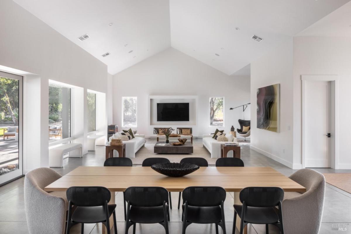 A dining area with a wooden table and black chairs leads to a living room with sofas and a TV.