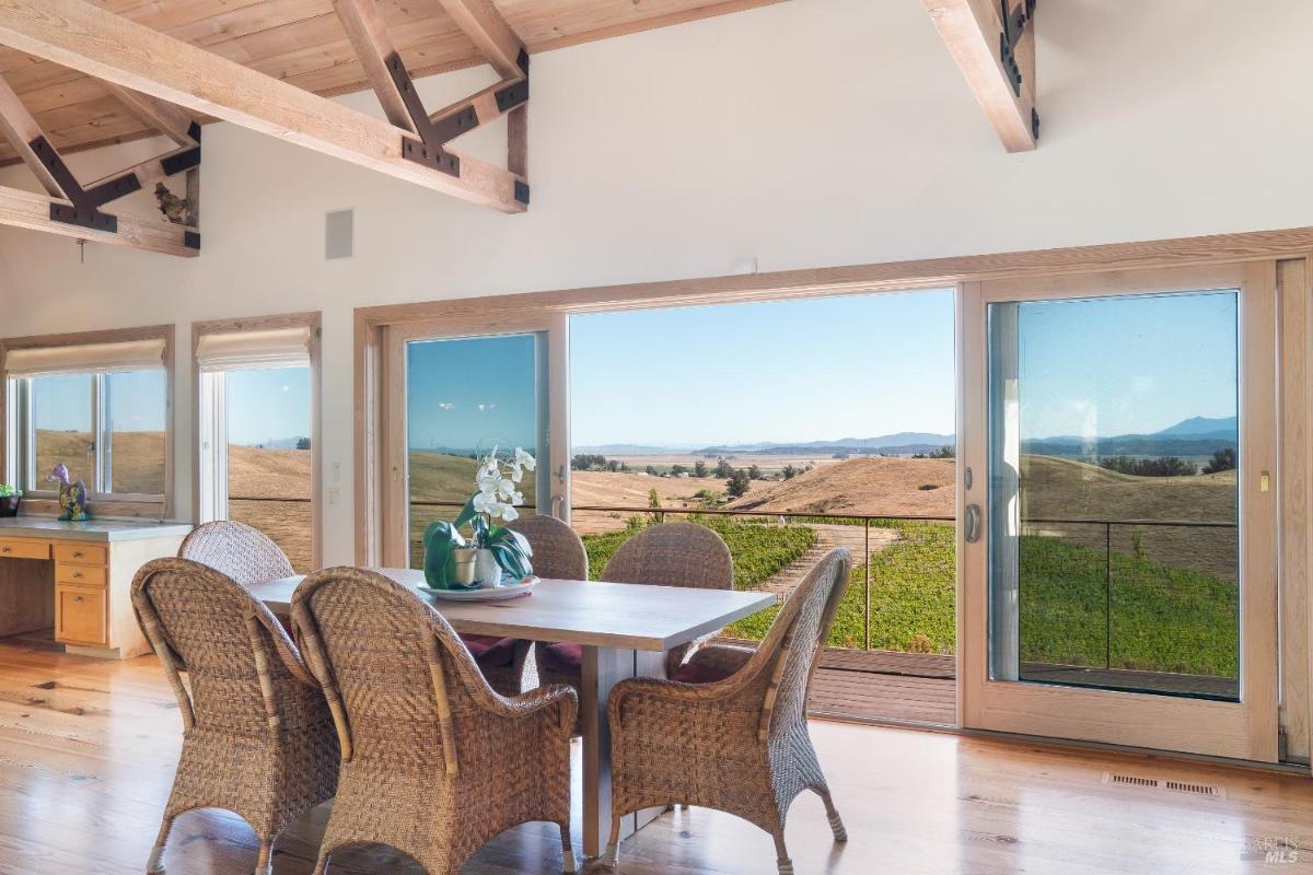 Dining area with wicker chairs, a wood table, and glass doors leading to a balcony overlooking vineyards.