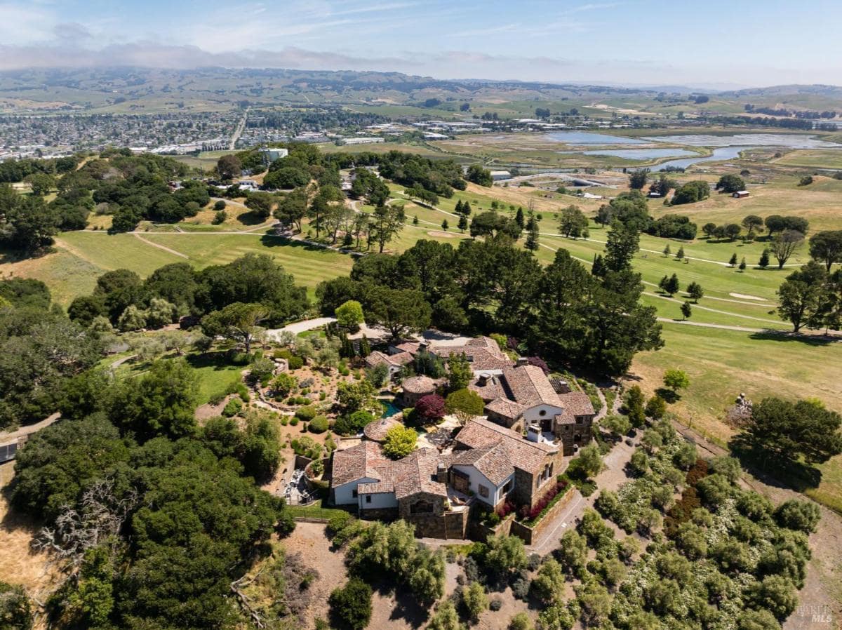 An aerial view of a property surrounded by trees and fields, with a cluster of buildings and a water body in the background.