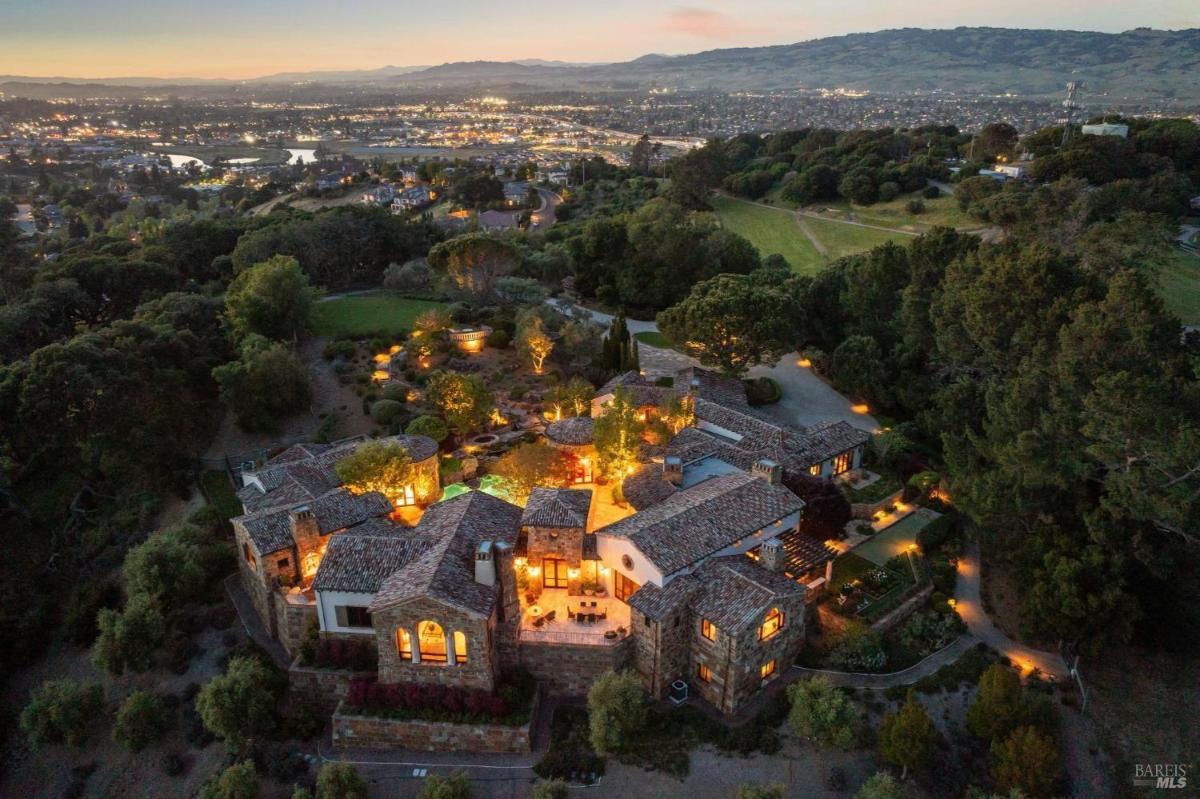 An aerial view of a well-lit property at dusk, featuring a cluster of buildings surrounded by trees and gardens, with a cityscape and hills in the background.