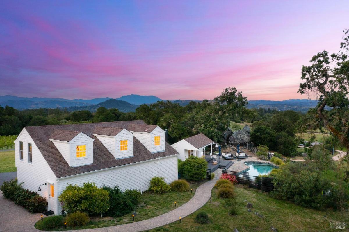 An aerial view of a white house with a pool and vineyard in the background. 