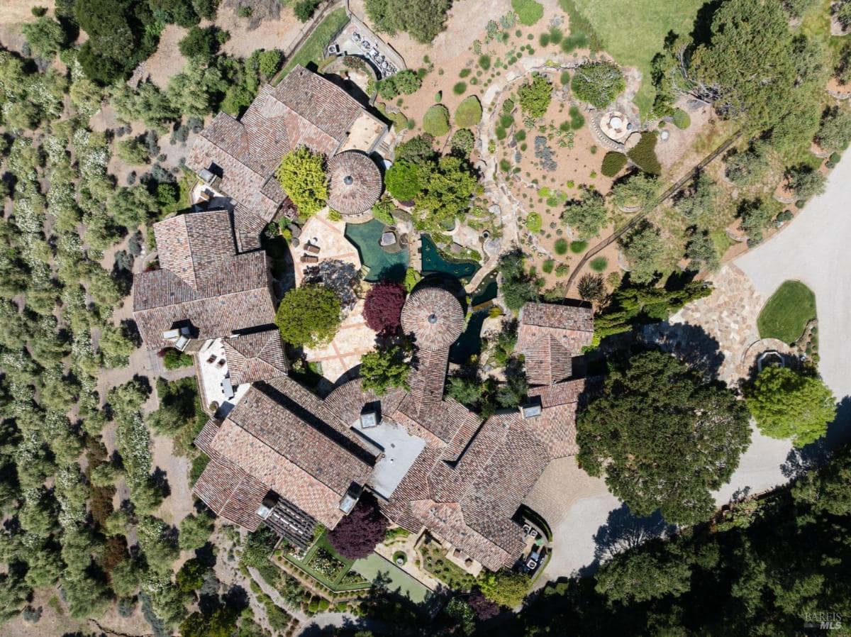 An aerial view of a residential house, featuring multiple buildings, gardens, and a pool, surrounded by trees and landscaped areas.