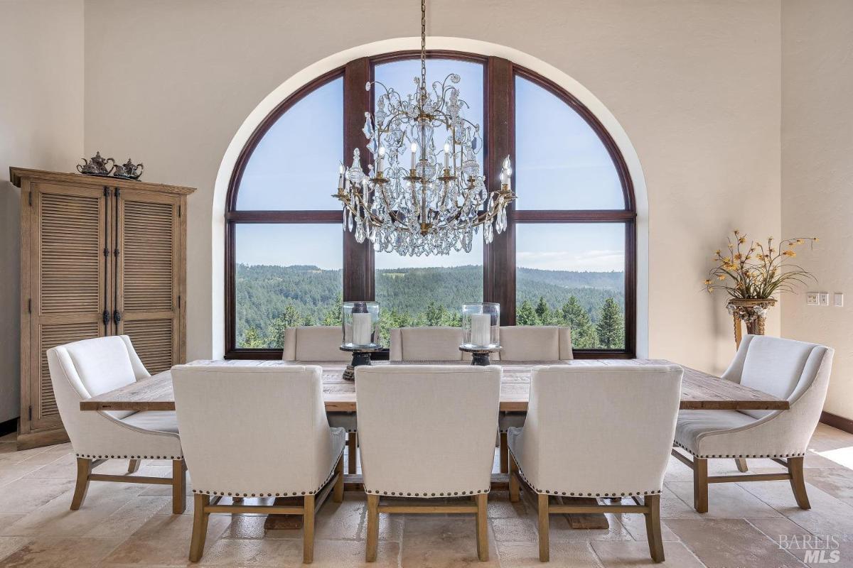 Dining area with a wooden table, white chairs, and a chandelier under an arched window.