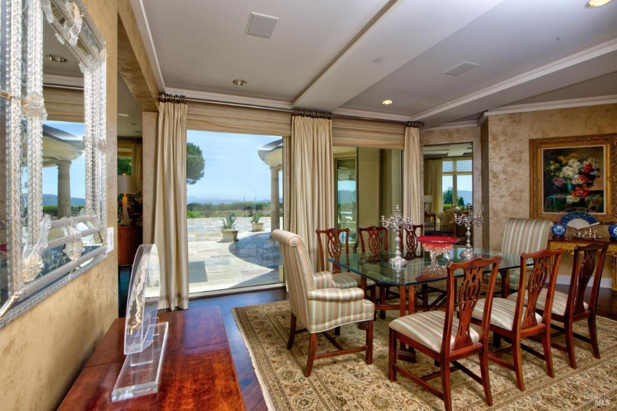 Formal dining area with glass table, upholstered chairs, and floor-to-ceiling windows leading to an outdoor patio and scenic views.