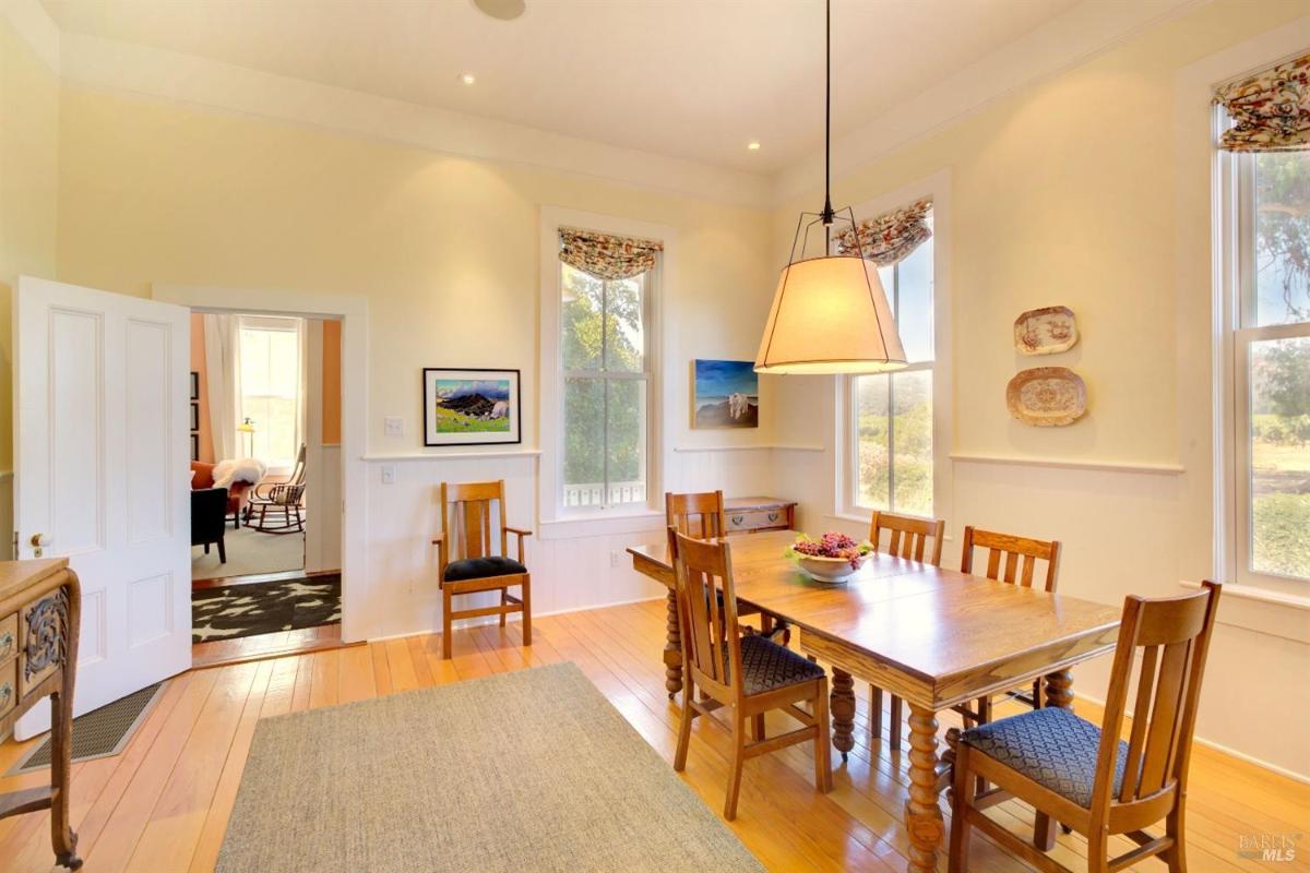 A dining room with white walls, large windows, a wooden dining table, chairs, and a hanging light fixture.