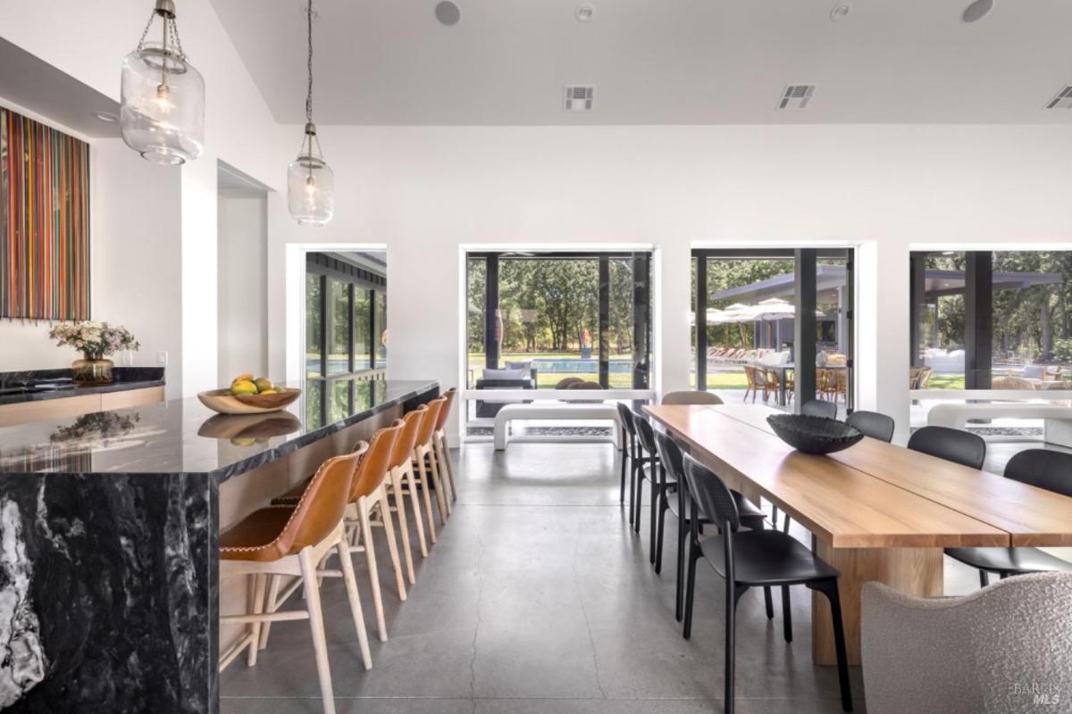 A kitchen bar with leather stools faces a wooden dining table surrounded by black chairs, with large windows showcasing an outdoor view.