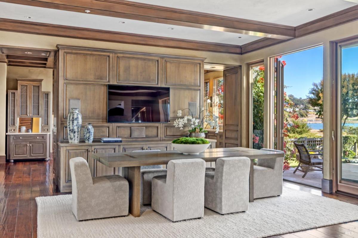 A dining area with a wooden table, four chairs, and a mounted television. Large windows show an outdoor view.