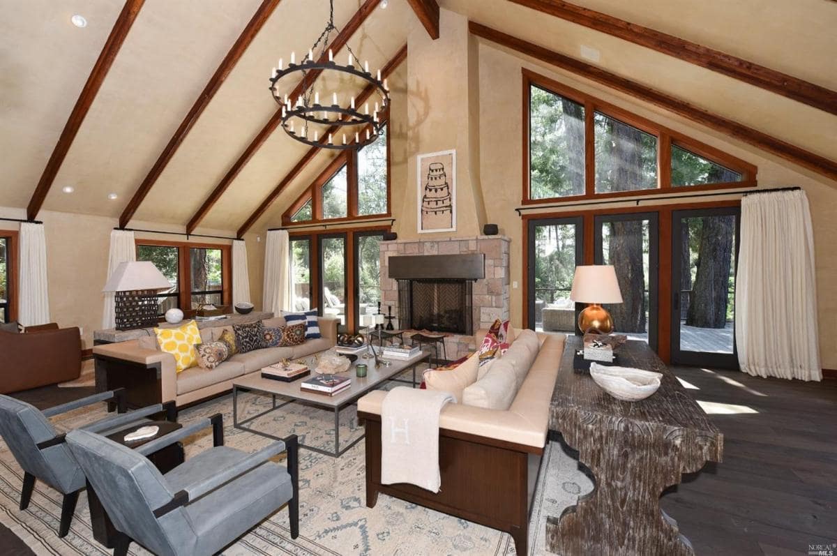 A living room with a stone fireplace, sofas, and forest views through large windows.