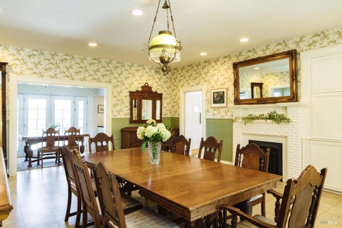 A dining room with a long wooden table and chairs, fireplace, and floral wallpaper.