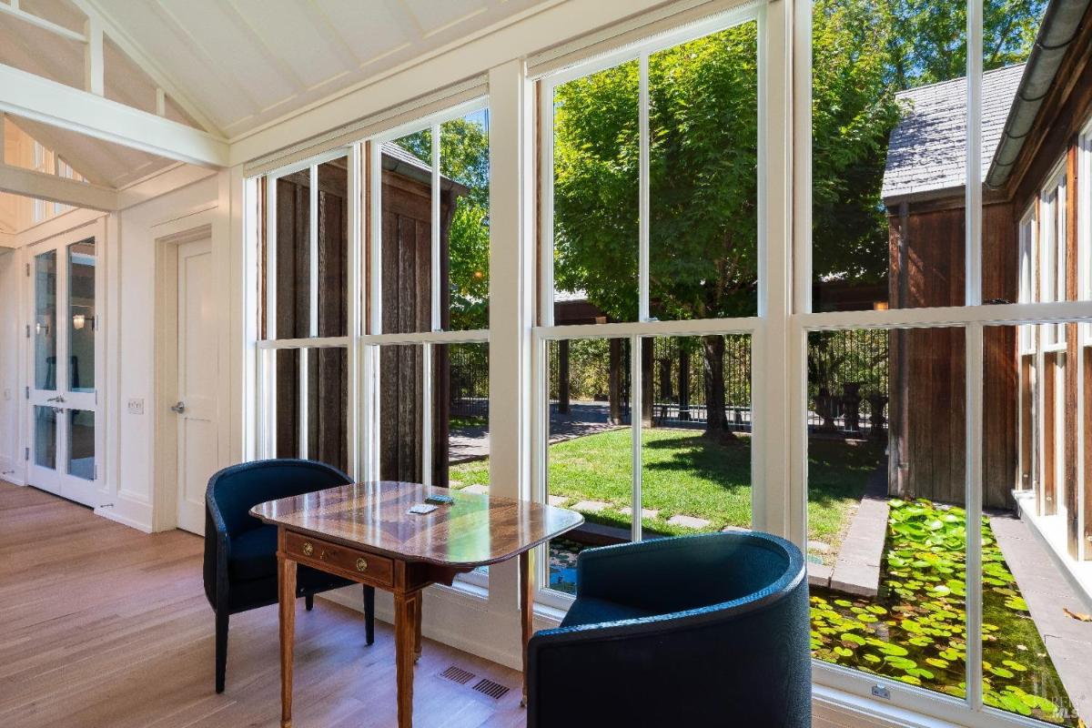 Indoor sitting area with a table, two chairs, and views of a garden and pond through windows.