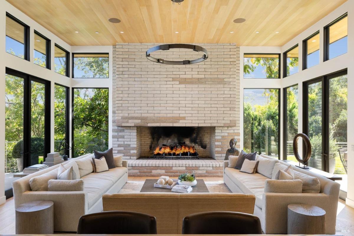 Close-up of a living room with a brick fireplace, a coffee table, sofas, and large windows providing natural light and garden views.