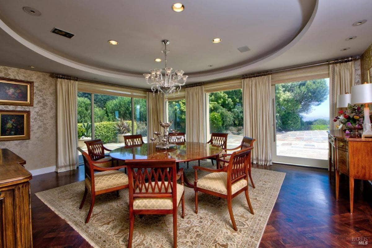 Circular dining room featuring a polished wood table, a chandelier, and wrap-around glass doors overlooking a manicured garden.