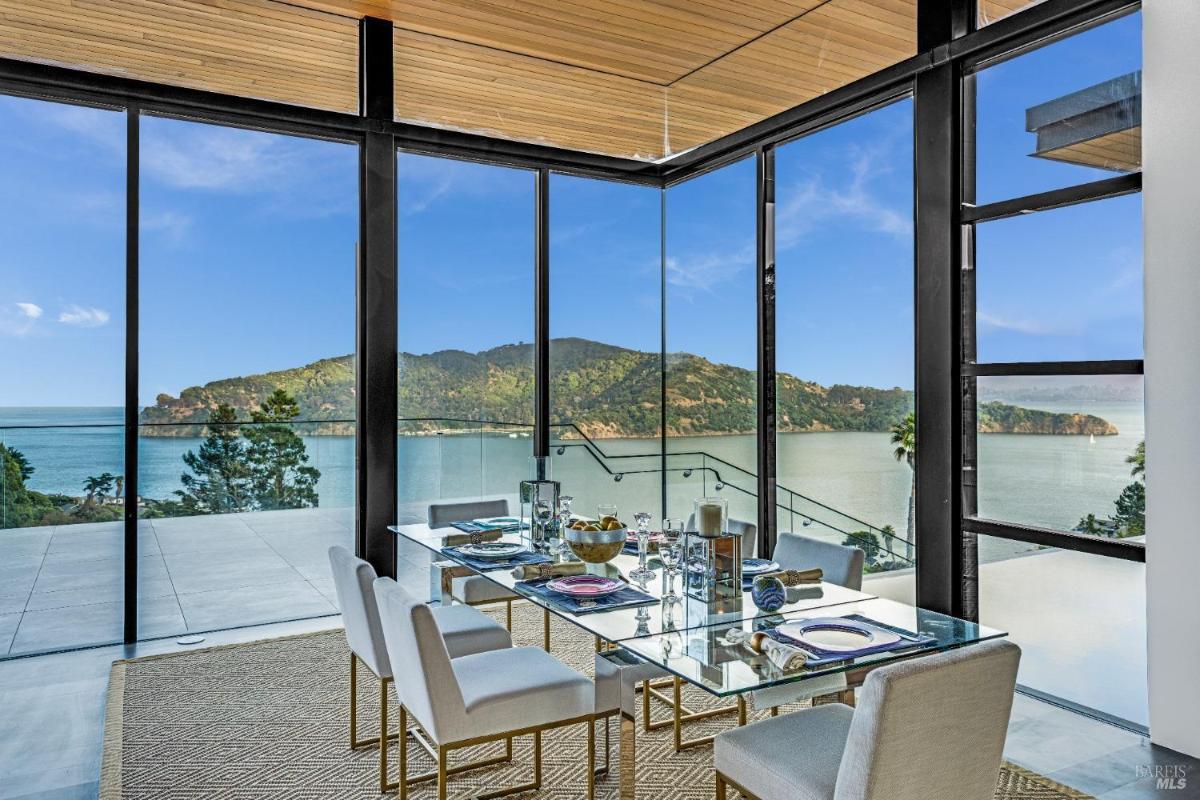 A dining area with glass dining table with chairs near floor-to-ceiling windows, and an overlooking view.