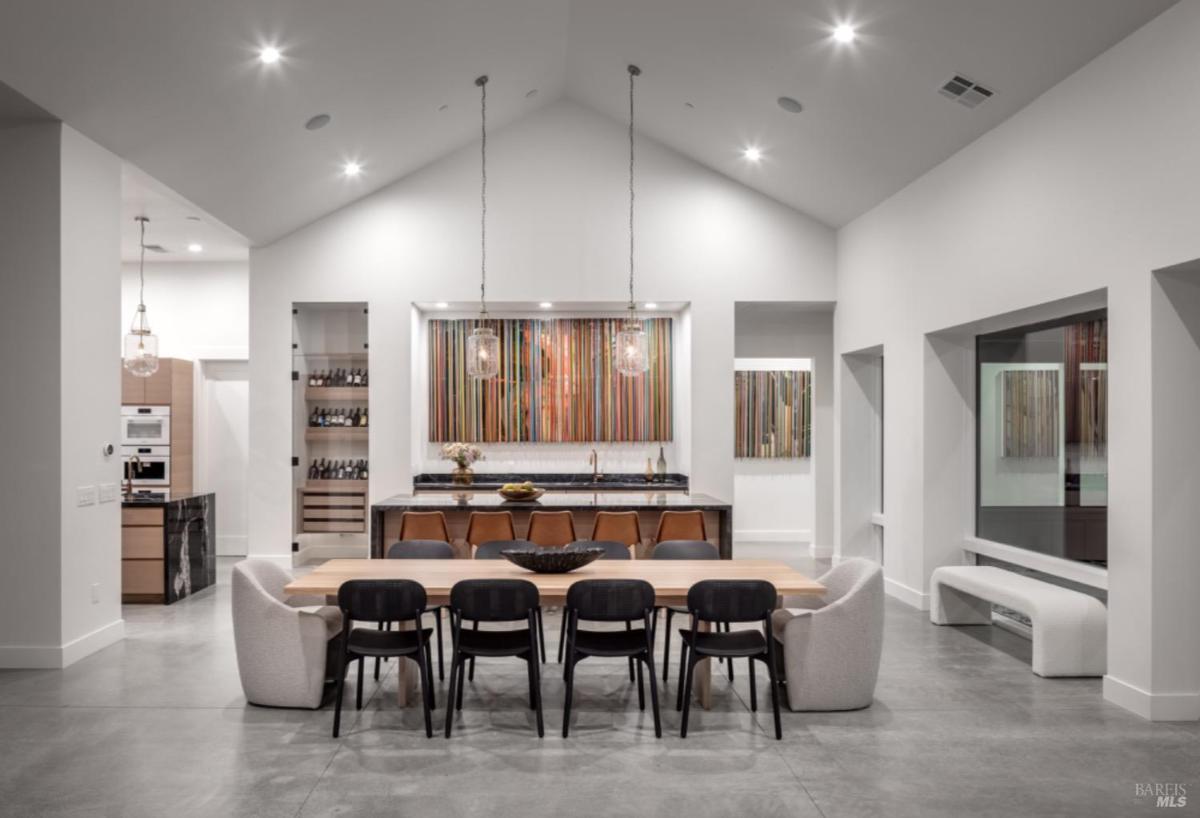 A modern kitchen with a marble island and bar seating connects to a wooden dining table with black chairs under vaulted ceilings.