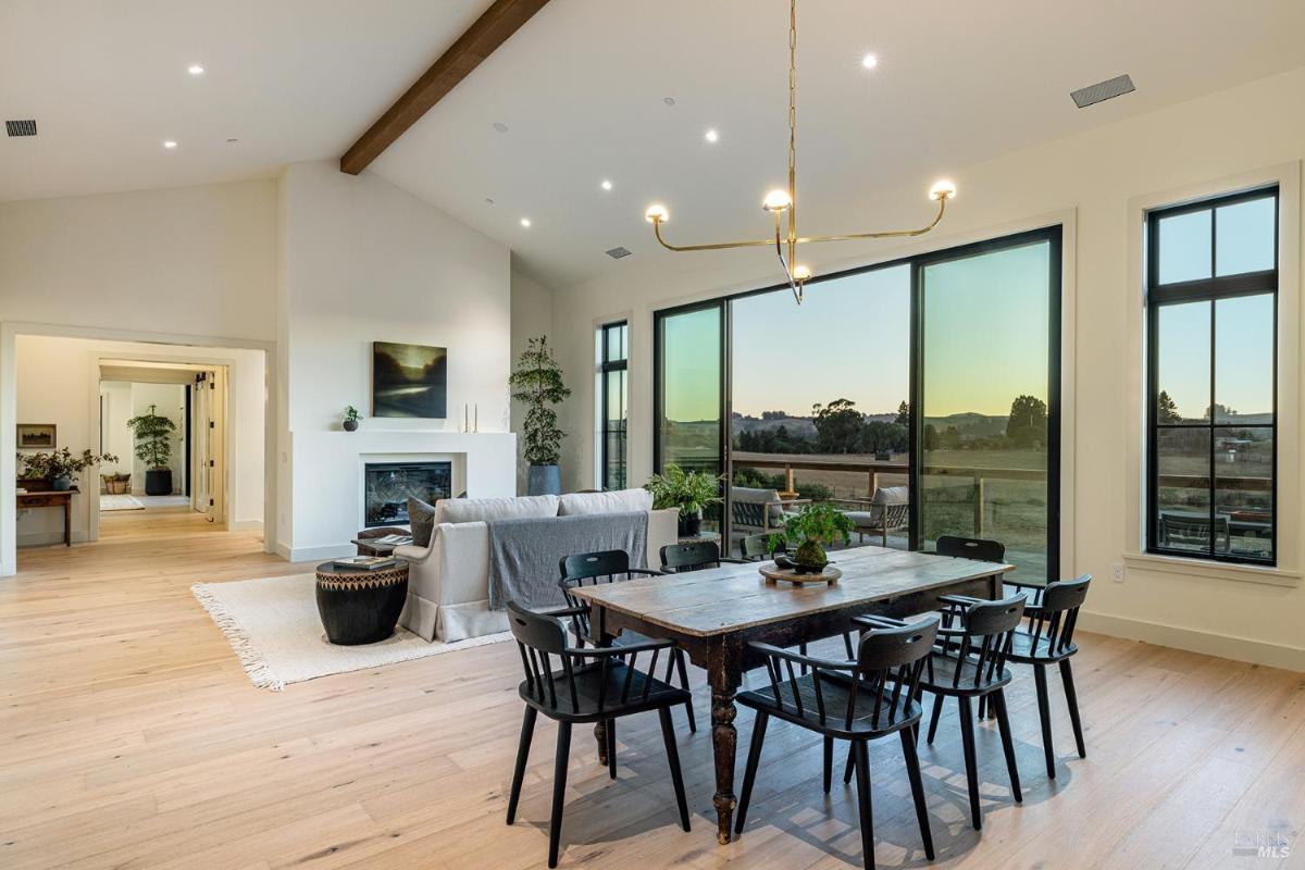 A dining area with a large table, chairs, a fireplace, and large windows showing an outdoor view.