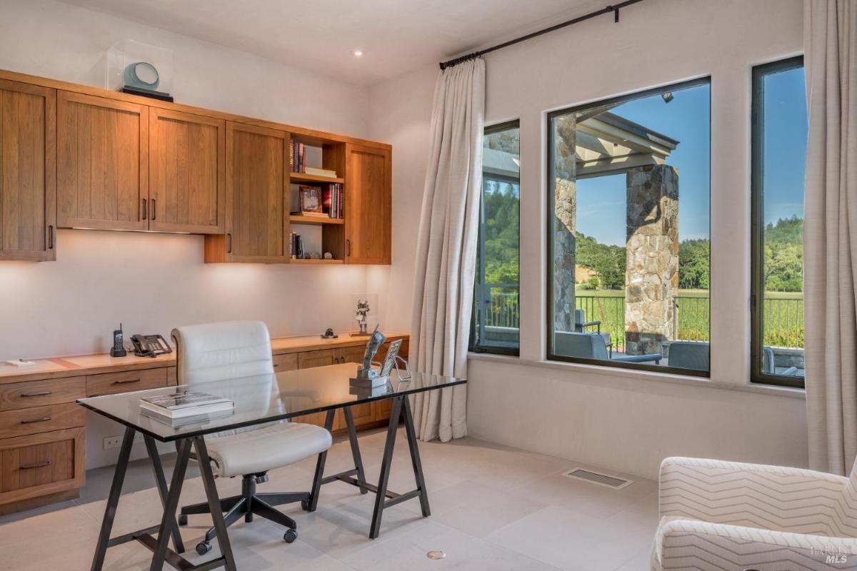 A home office with a desk, chair, bookshelves, and large windows overlooking an outdoor area.