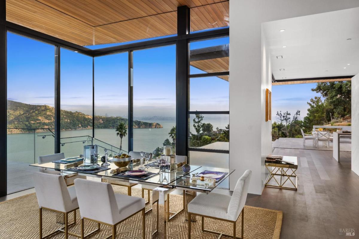 A dining room with a glass table, white chairs, and large windows overlooking a bay.