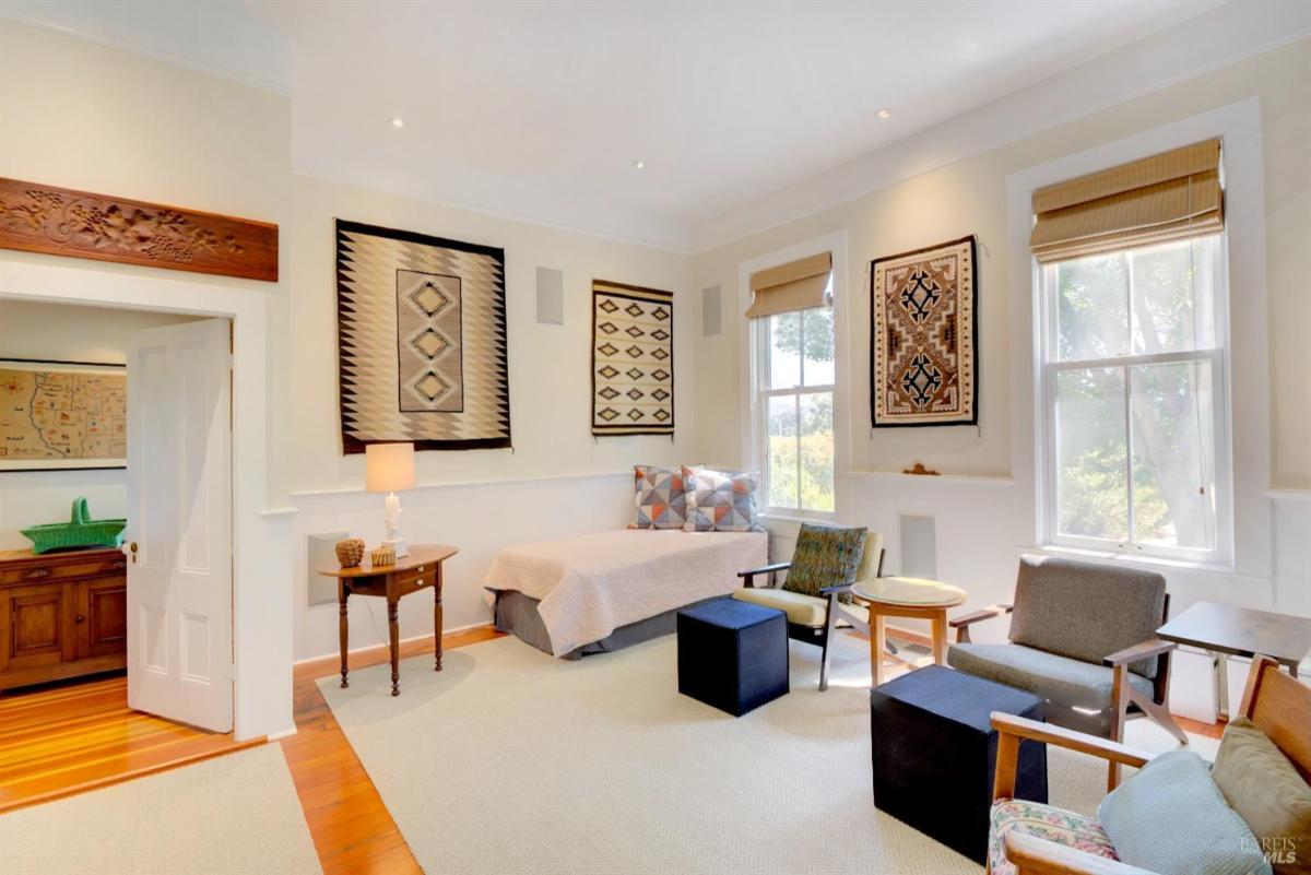 A family room with patterned wall hangings, chairs, a small bed, and a side table under large windows.