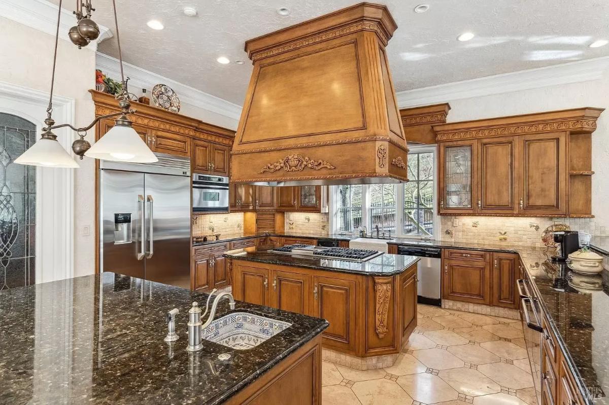 A kitchen with granite countertops, a large wooden hood, and an island with bar seating.