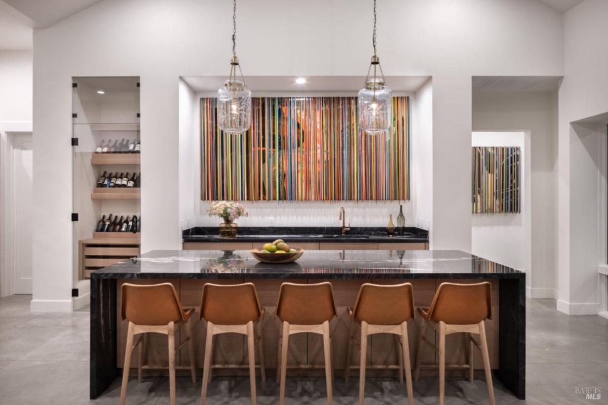 A modern kitchen features a black marble island with brown leather barstools, pendant lights, and a built-in wine display.