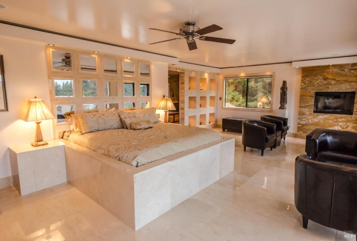 A contemporary bedroom with a platform bed, large windows, modern armchairs, and a stylish fireplace.