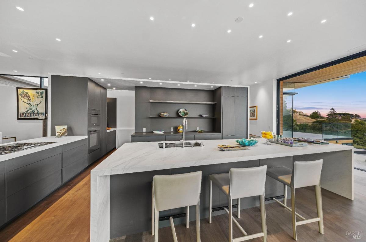 A modern kitchen with a large island, bar seating, and hillside views.