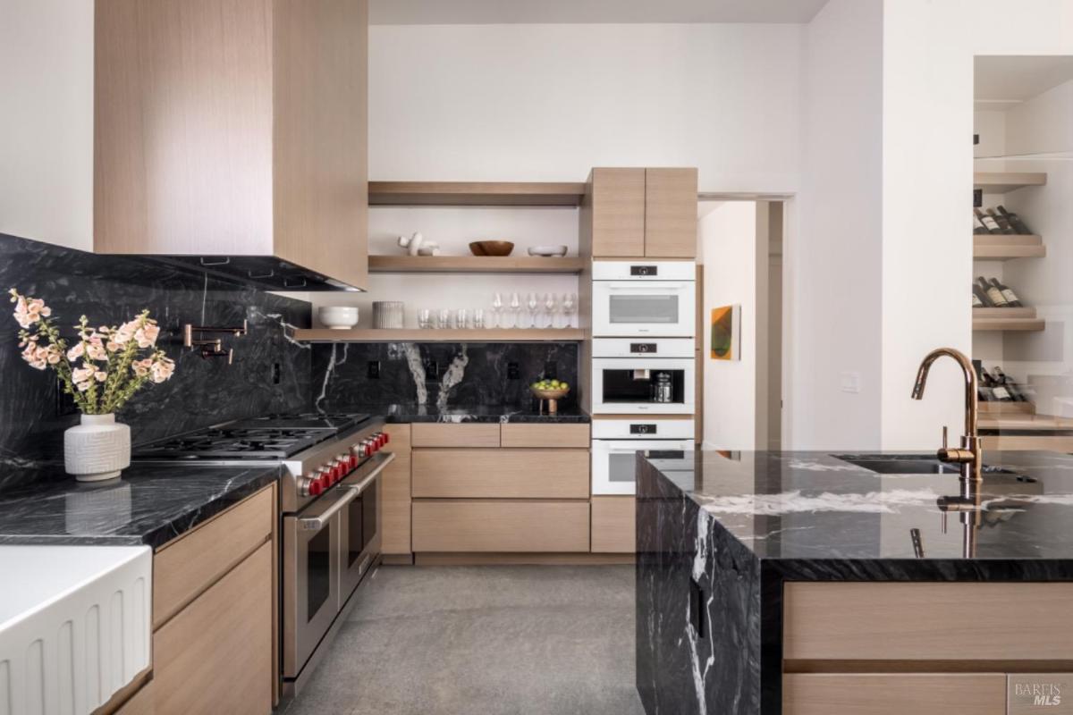 A kitchen features a black marble backsplash and island, light wood cabinetry, stainless steel appliances, and open shelving.