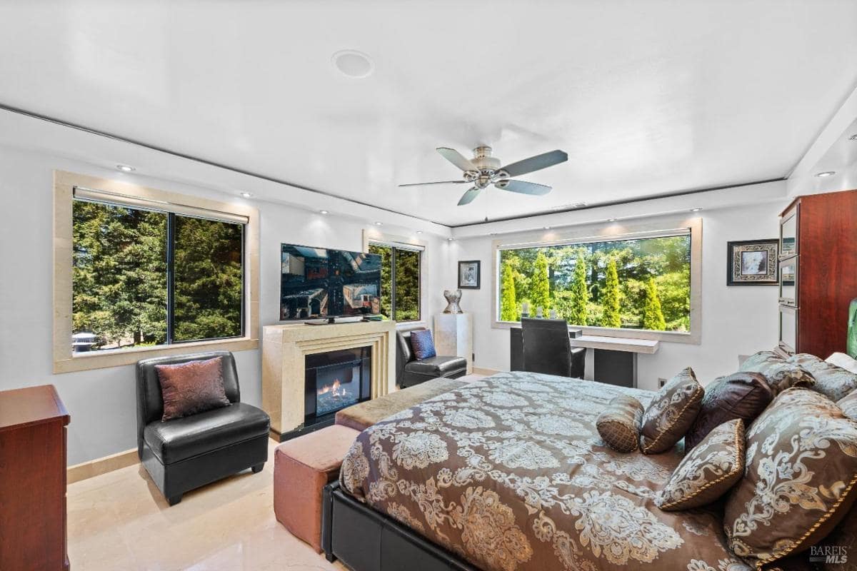 A bedroom with a king-size bed, two armchairs, a television mounted on the wall, a fireplace, and large windows showing greenery outside.