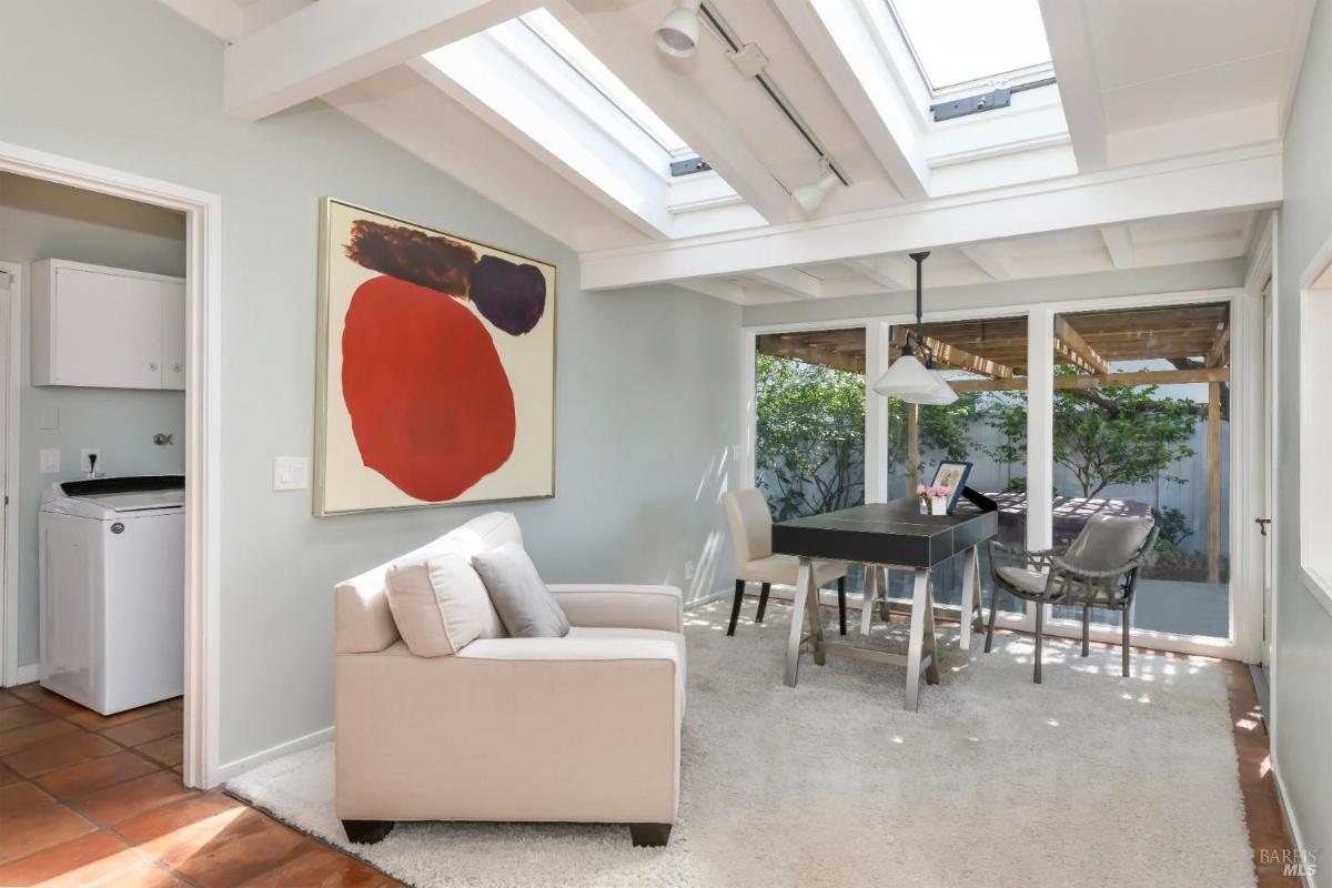 A home office with a skylight, armchair, and desk, adjacent to a glass door opening to a pergola-covered outdoor space.