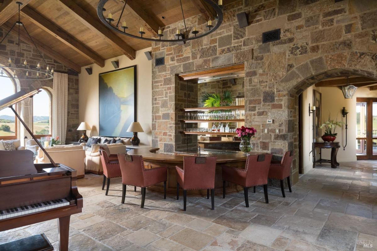 A bar area with a stone wall, a large round table surrounded by chairs, a grand piano nearby, and large windows showcasing a view of the outdoors.