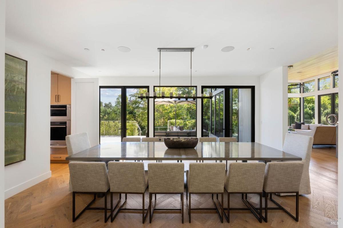 A dining area with a long table, chairs, and large windows providing views of trees and an outdoor patio.