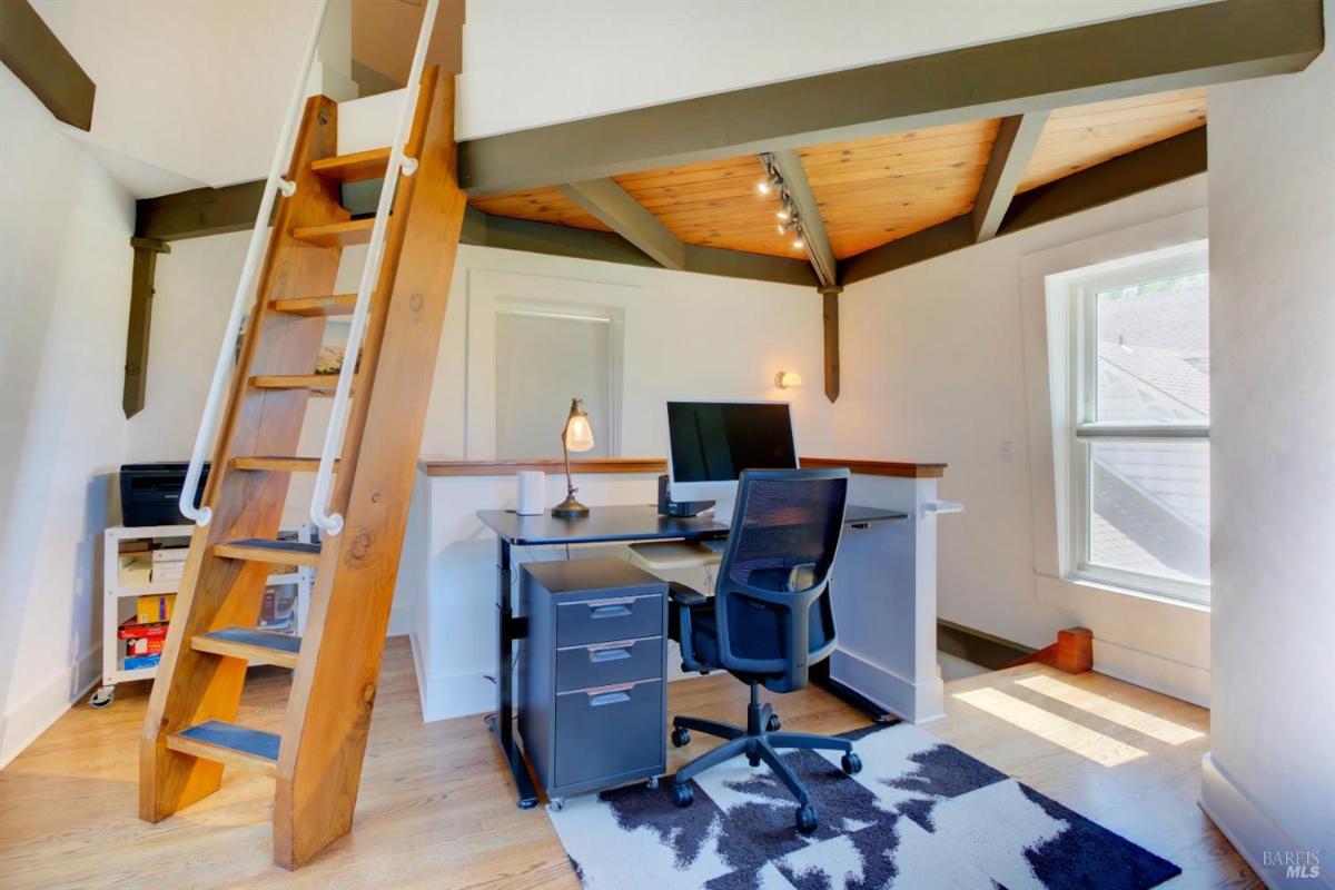 A home office with a wooden ladder leading to a loft, a desk with a monitor, and a window letting in natural light.