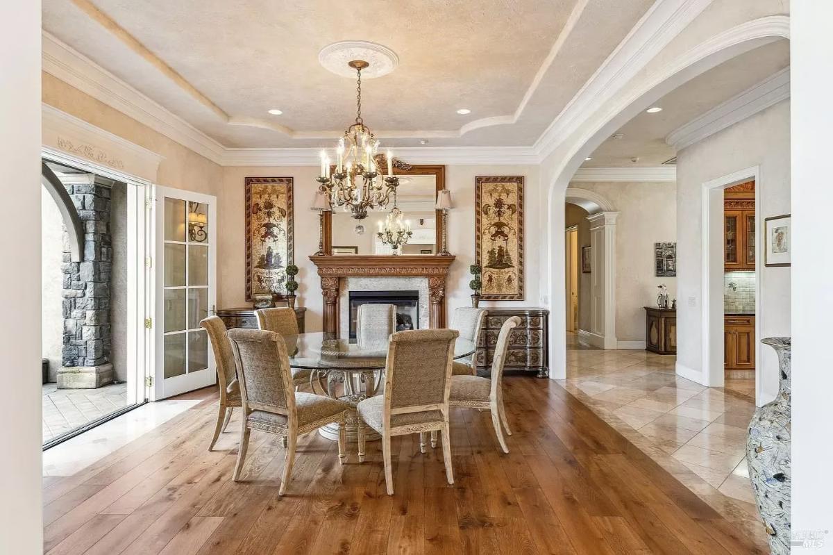 Dining area with a round glass table, six chairs, a decorative chandelier, and a fireplace with a wood mantel.