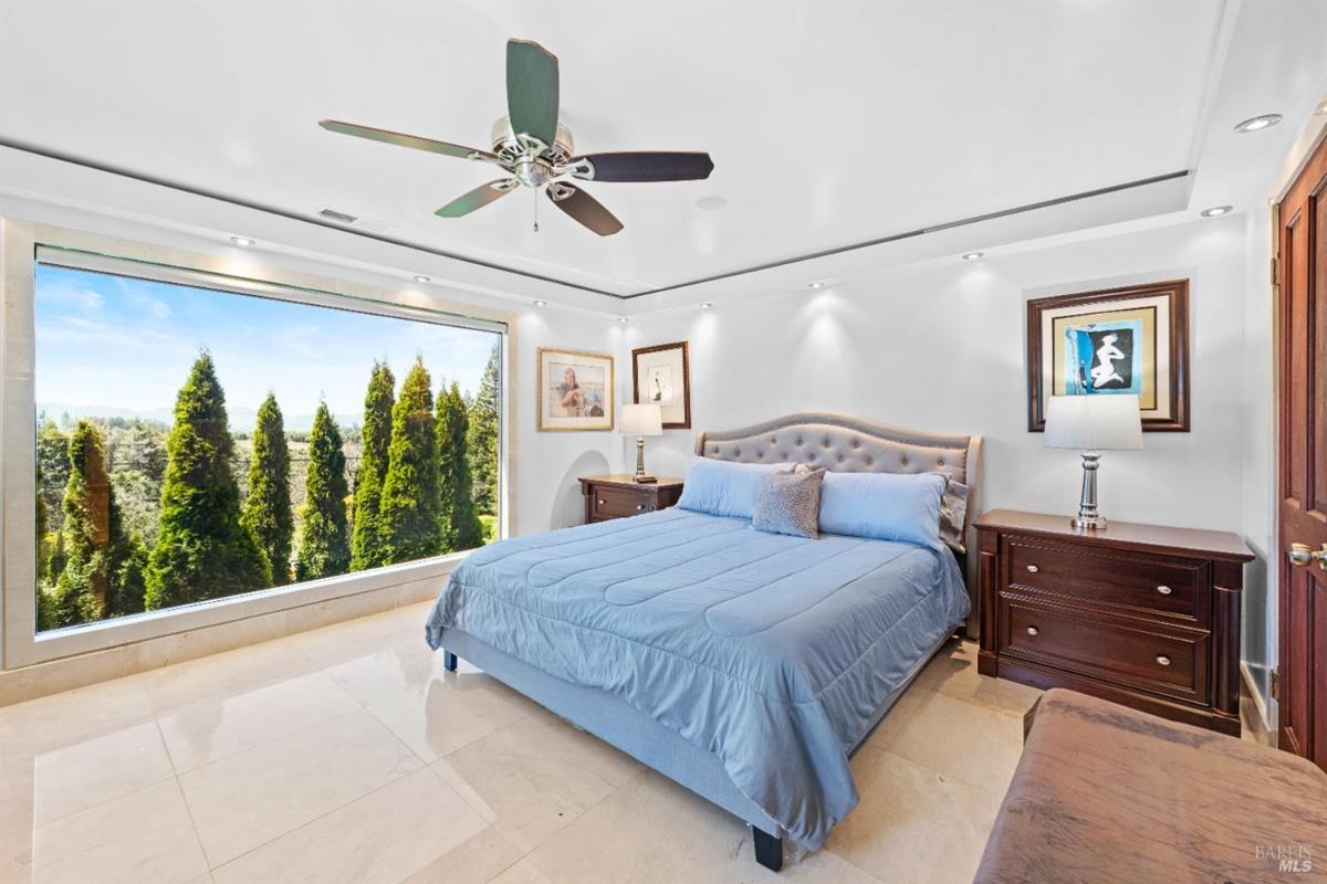 A bedroom with a queen-size bed, two nightstands, a ceiling fan, and a large window displaying a view of trees outside.