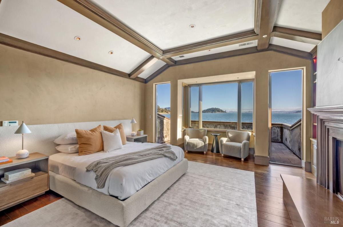 A bedroom with a large bed, two chairs, and a view of the water through large windows. The room has a neutral color scheme and wooden accents.