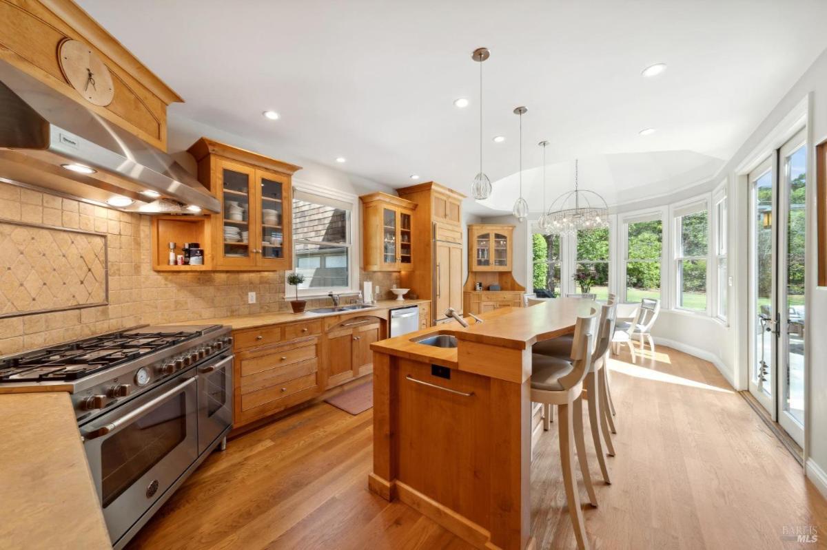 A kitchen area featuring stainless steel appliances, pendant lighting, and a dining nook with large windows.