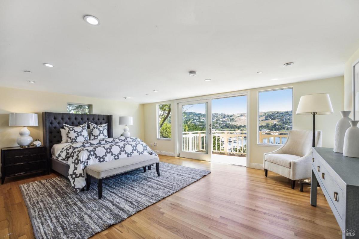 Bedroom with a bed, chairs, and a balcony offering views of water and hills.