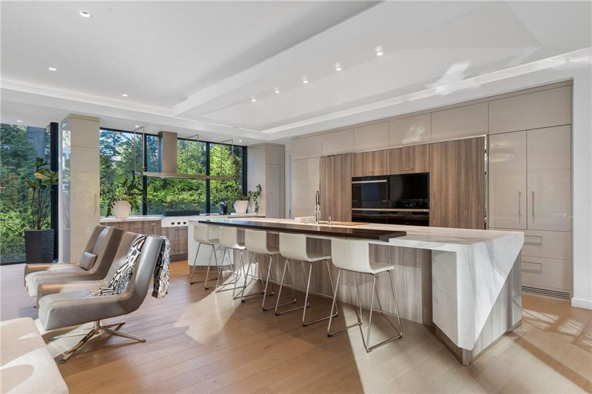 Kitchen features an oversized waterfall island, high-end appliances, and expansive windows framing lush greenery.