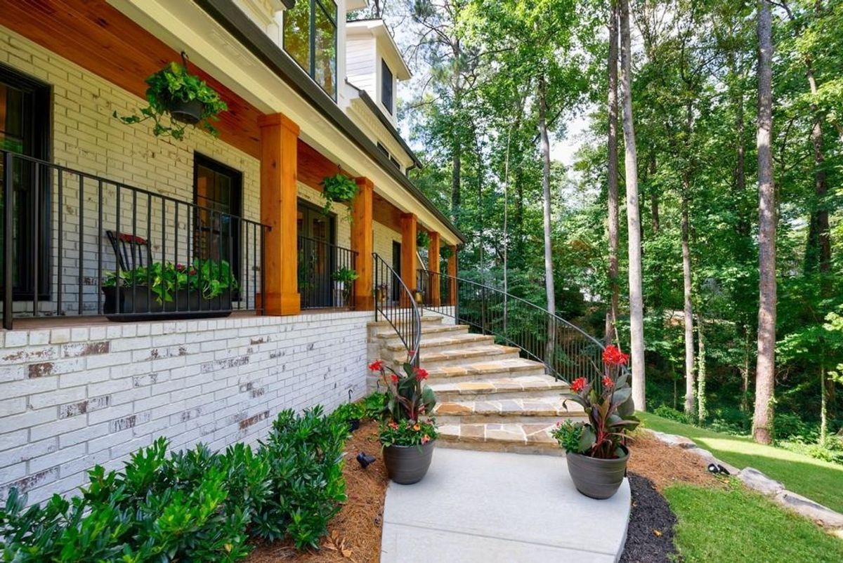 Elegant stone steps lead to a welcoming porch adorned with wood beams, hanging plants, and a serene forest backdrop.