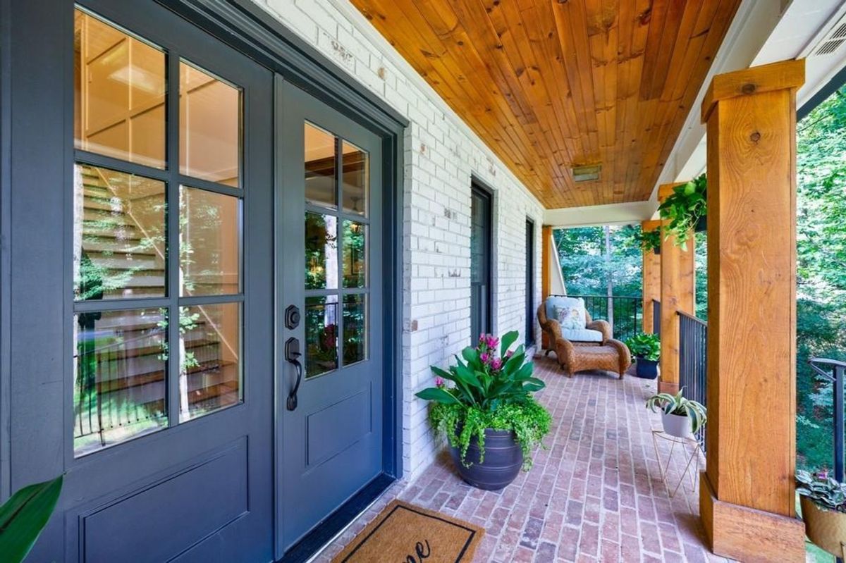 Brick flooring and wood-beamed ceilings create a warm and inviting front porch space, complemented by vibrant plants and cozy seating.