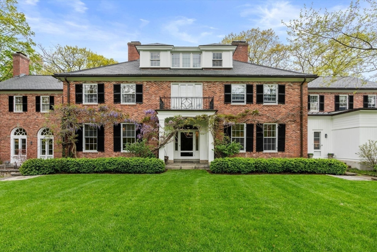 A stately red brick Colonial home with black shutters and lush greenery radiates timeless elegance and charm.