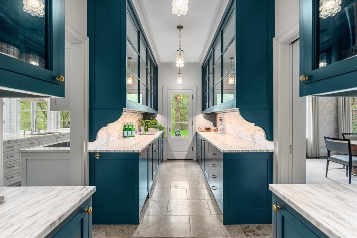 A striking butler’s pantry showcases deep teal cabinetry, marble countertops, and glass-front displays illuminated by elegant pendant lighting.