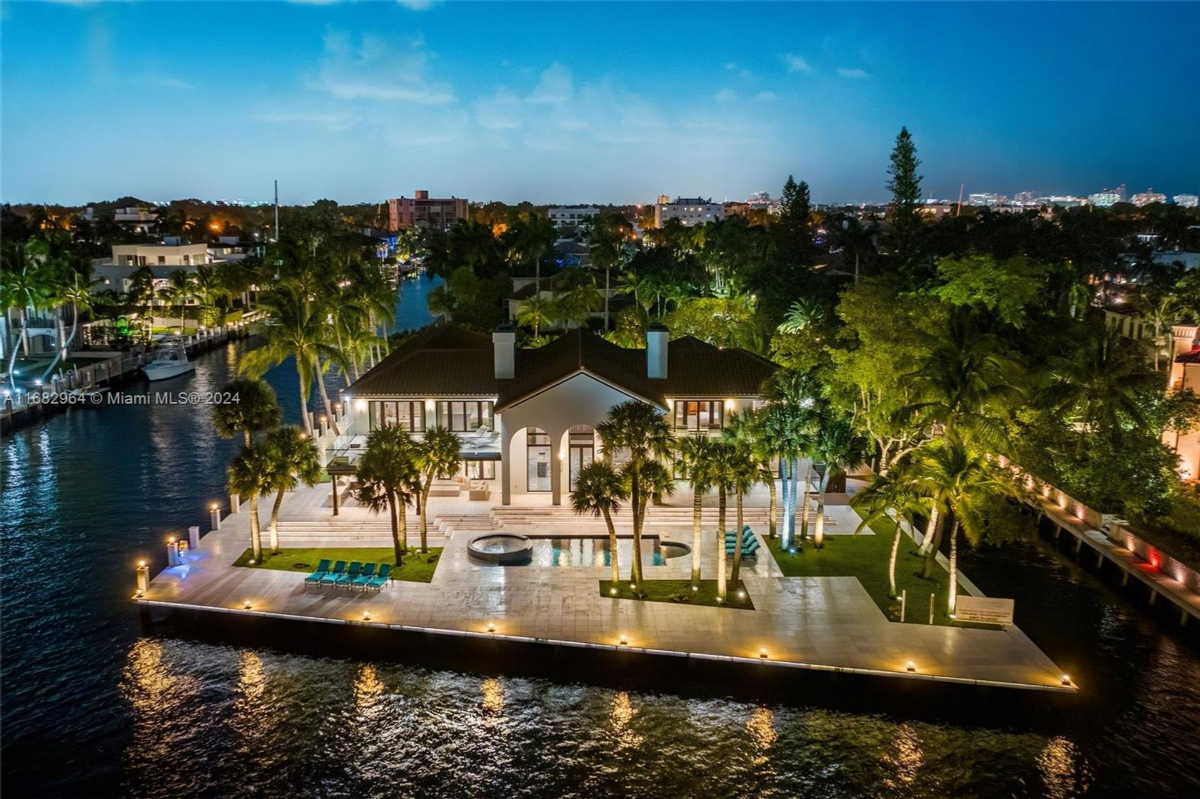 An illuminated waterfront mansion shines against the tranquil night, framed by palm trees and shimmering waters.