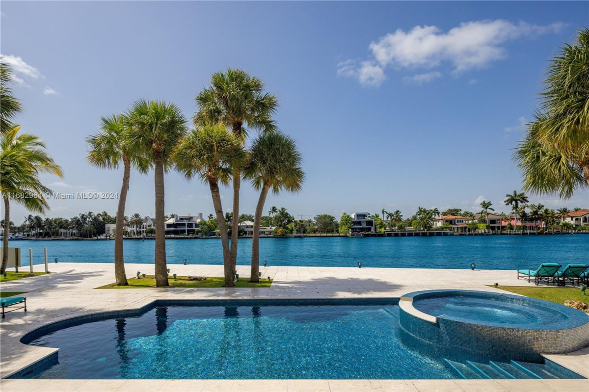 Elegant infinity-edge pool and spa overlooking a pristine waterfront lined with palm trees and luxurious residences.