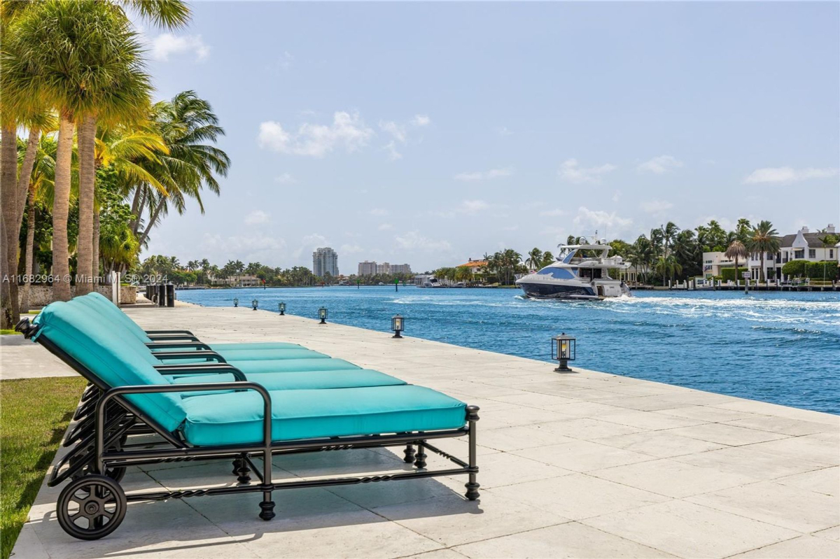 Lounge chairs with vibrant cushions arranged alongside a waterfront terrace, offering serene views of passing yachts and palm-lined shores.